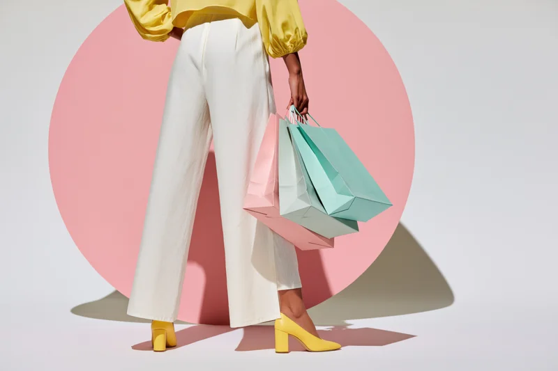 Woman from waist down holding pastel shopping bags in front of pink circular backdrop