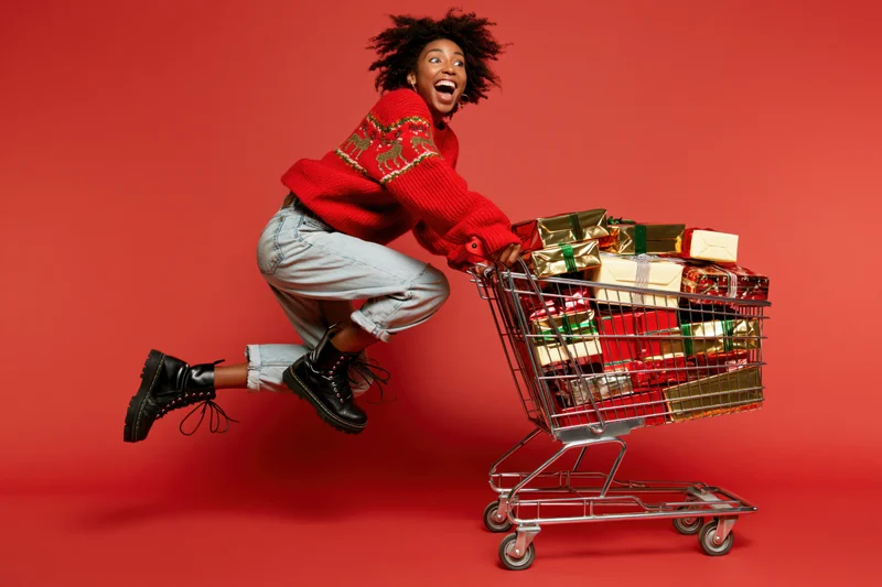 Young black woman mid-jump pushing a metal shopping cart filled with wrapped gifts against a red background