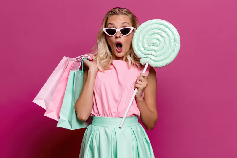 Blonde woman with open-mouthed surprised expression holding oversized lollipop and shopping bags against pink background