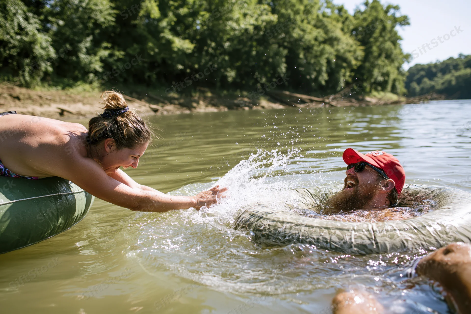Woman leaning from green inflatable tube splashing water at man in another tube on river