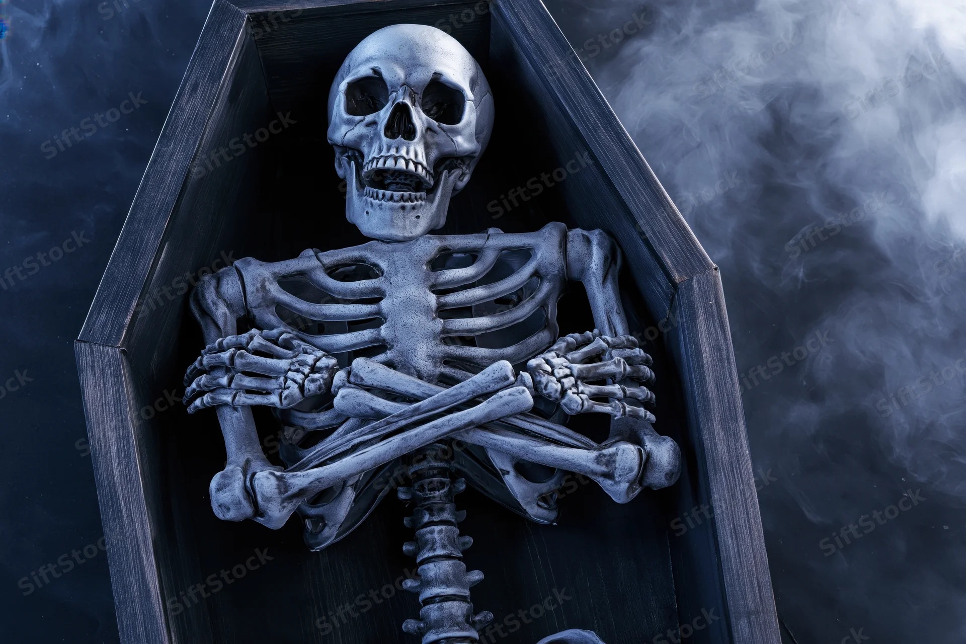 Skeleton with crossed arms lying inside a wooden coffin surrounded by smoke