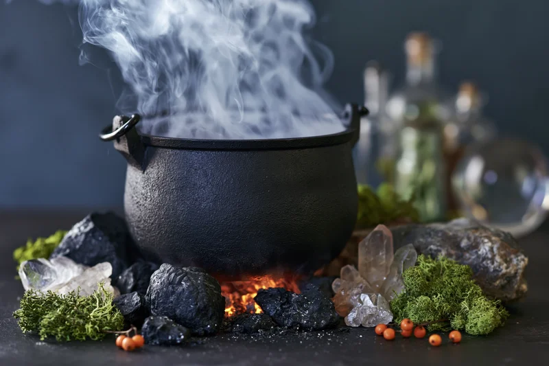 Black cast iron cauldron billowing smoke over fire surrounded by crystals moss and berries on dark surface