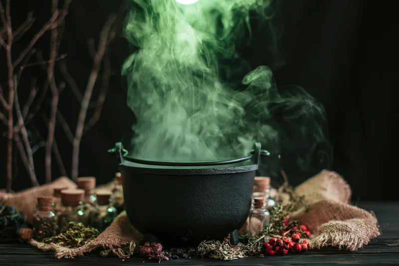 Black cauldron emitting swirling green smoke on dark wooden table surrounded by herbs, berries, glass bottles and burlap cloth