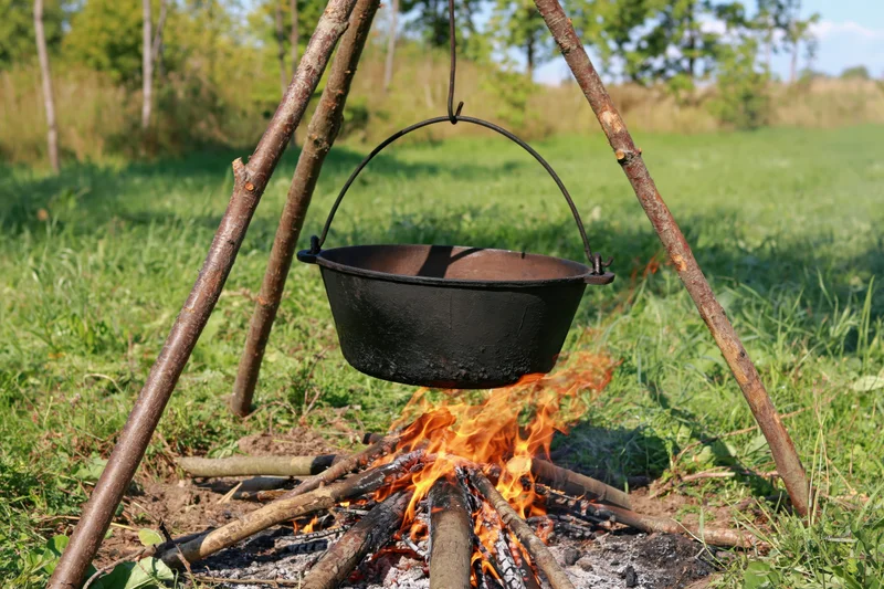 Blackened cast iron pot hanging over a campfire on a wooden tripod in a grassy field