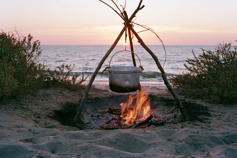 Metal pot hanging over a campfire on a sandy beach at sunset by the ocean