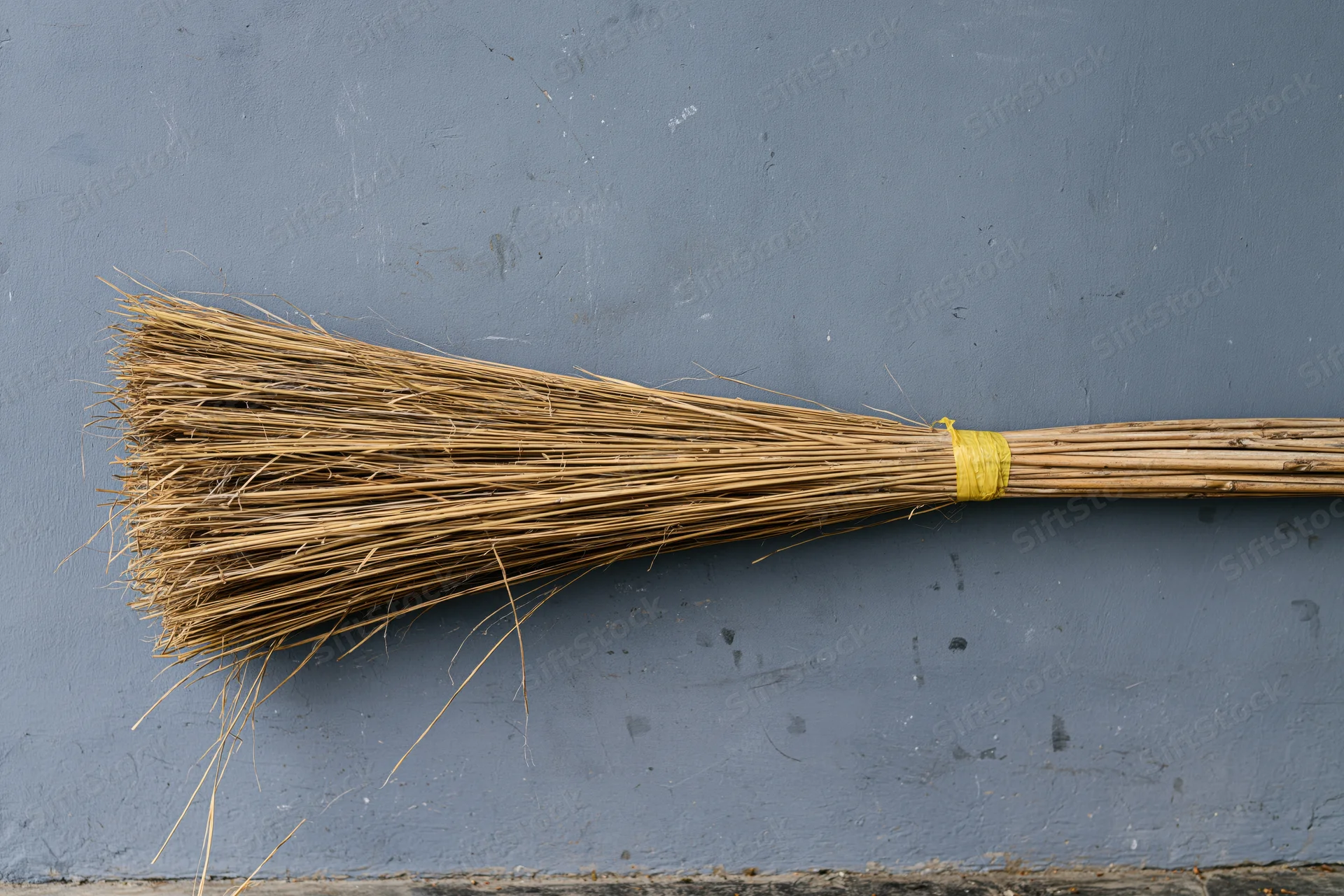 A straw broom lying flat against a gray concrete wall