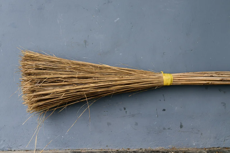 A straw broom lying flat against a gray concrete wall