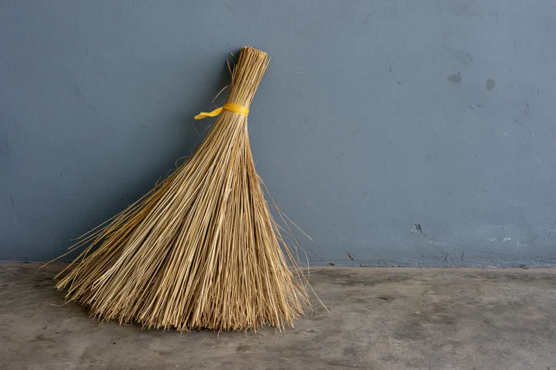 A straw broom leaning against a gray wall on a concrete floor