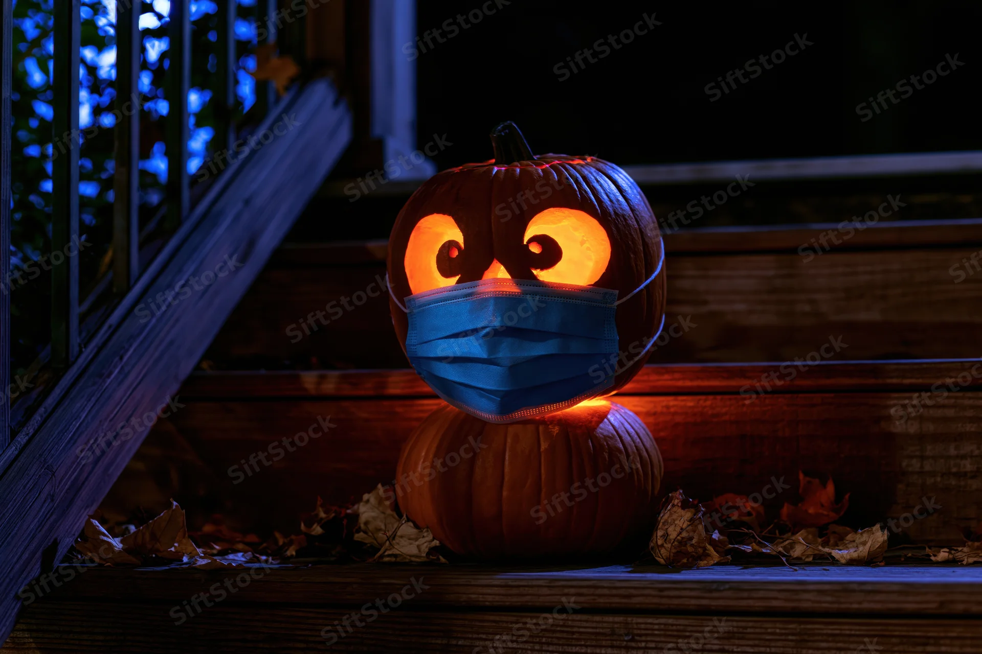 Carved jack-o-lantern wearing blue surgical mask glowing on wooden porch steps at night