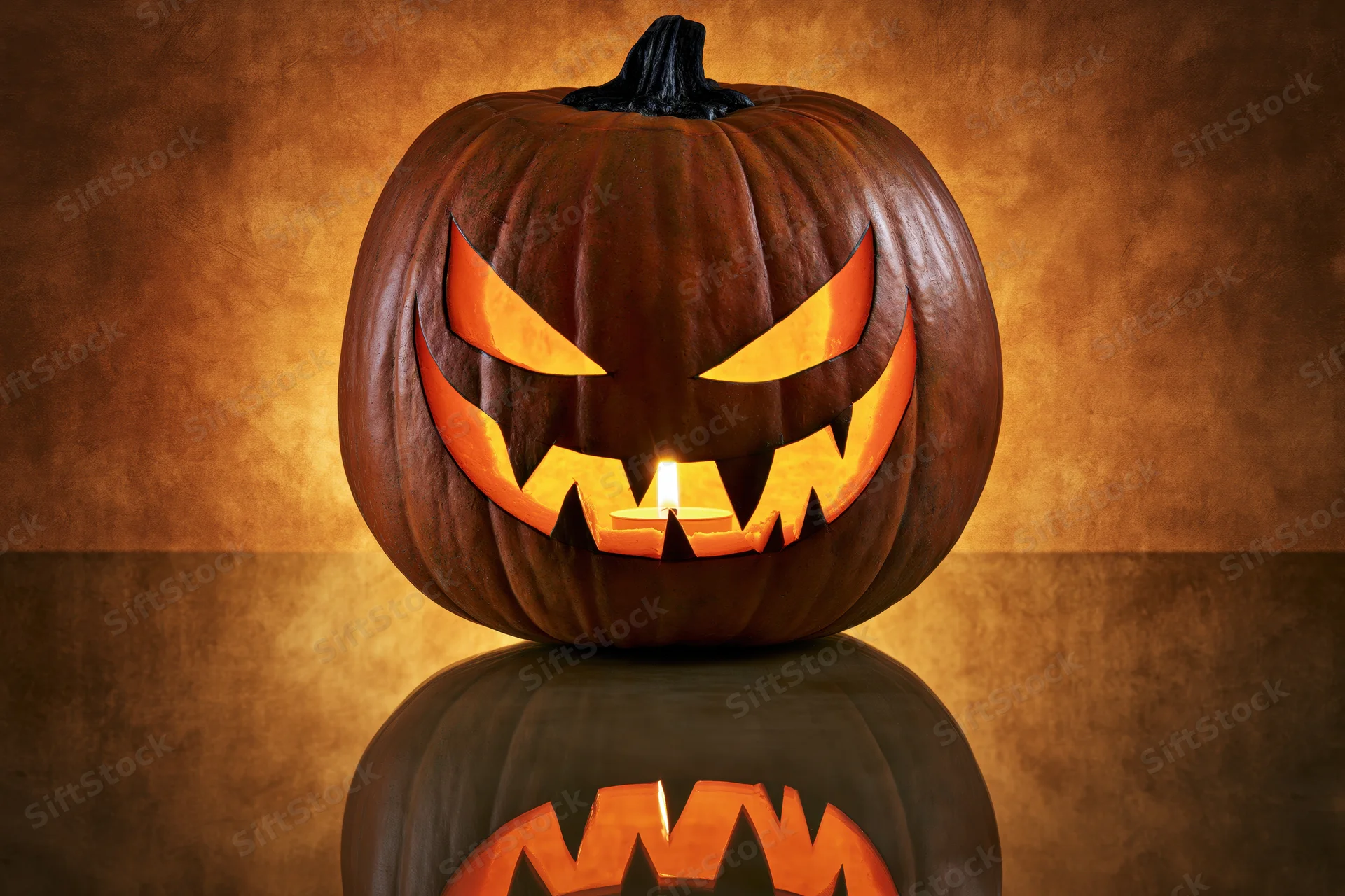 Carved pumpkin with menacing grin lit by candle on reflective surface