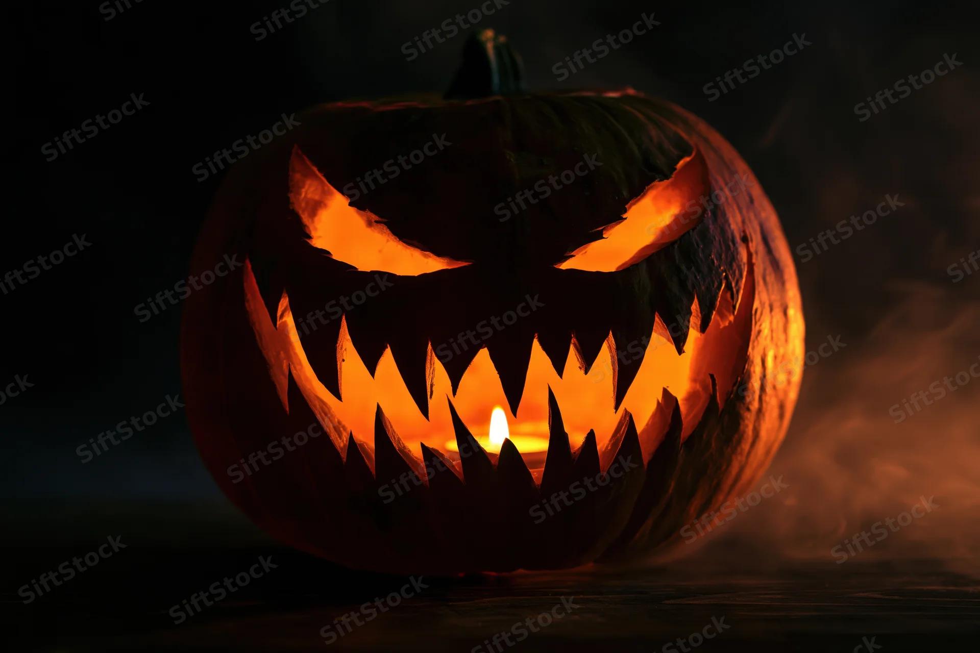 Carved pumpkin with jagged teeth and glowing eyes lit by candle in dark smoky setting