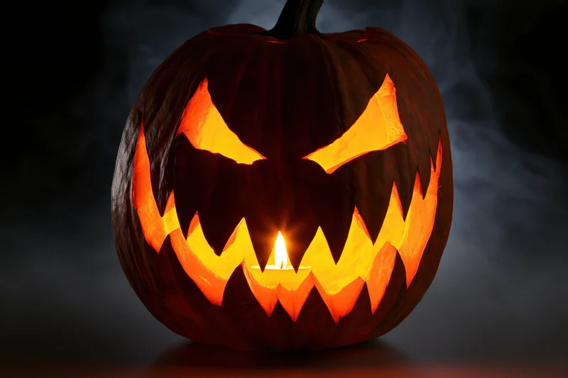 Carved pumpkin with jagged teeth and glowing eyes lit by candle in dark smoky setting