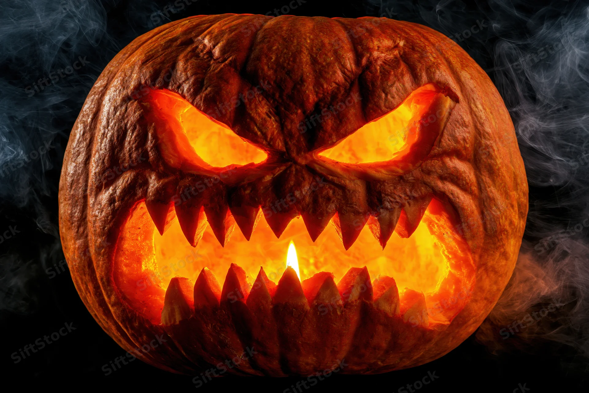 Carved pumpkin with glowing eyes and sharp teeth lit by candle in dark smoky setting