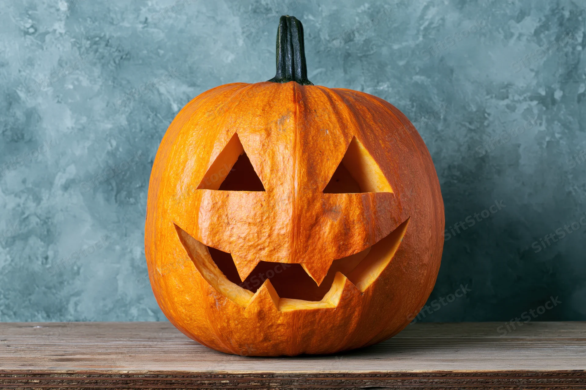 Carved pumpkin with triangular eyes and jagged grin on wooden table against textured blue-gray background