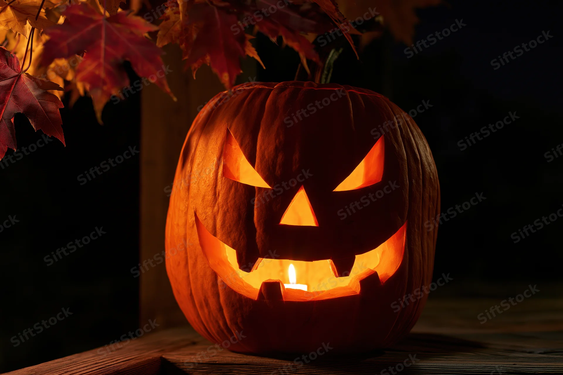 Carved pumpkin with glowing candle face on wooden surface under autumn leaves at night