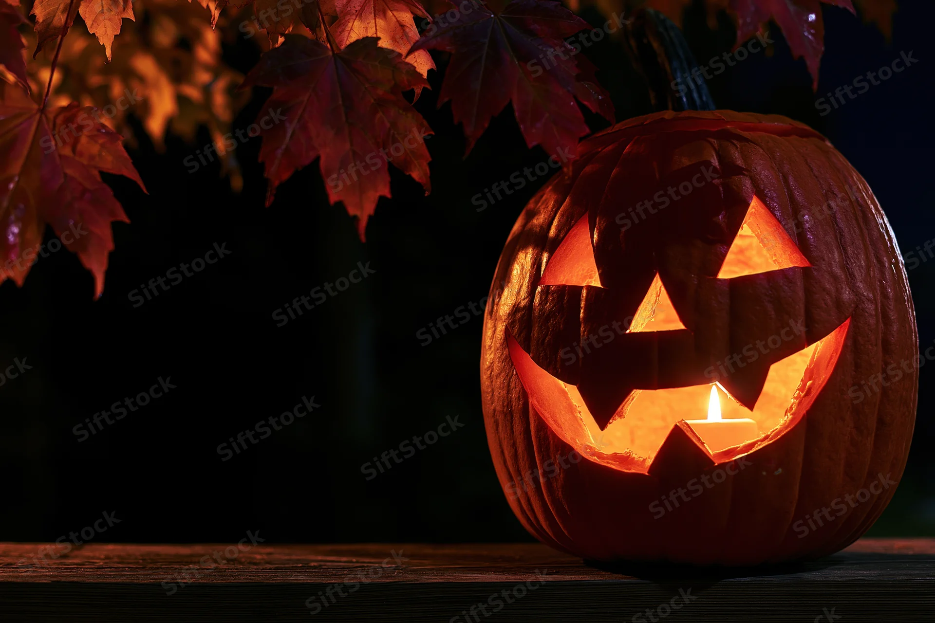 Carved pumpkin glowing with candlelight on wooden surface under autumn leaves at night