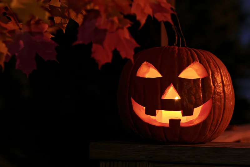 Carved pumpkin glowing with candlelight on wooden surface at night surrounded by autumn leaves