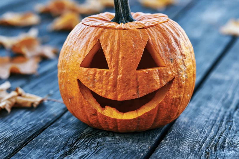 Carved jack-o-lantern with smiling face on dark wooden table surrounded by dry autumn leaves