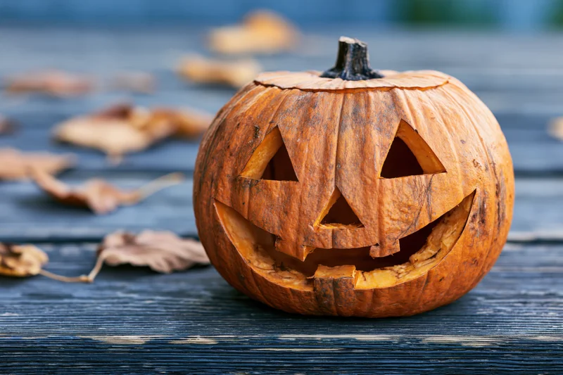 Carved pumpkin with smiling triangular eyes on a dark wooden table surrounded by autumn leaves