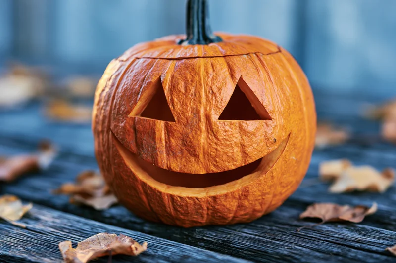 Carved jack-o-lantern with smiling triangular eyes on a dark wooden table surrounded by dried autumn leaves