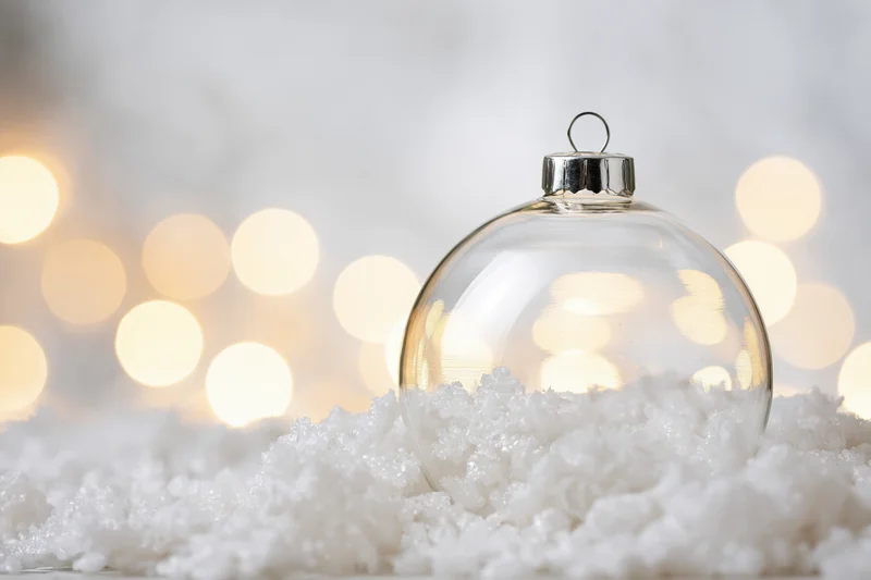 Clear glass christmas ornament resting in artificial snow with blurred warm lights behind