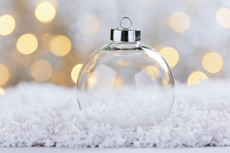 Clear glass christmas ornament resting on snow with blurred golden lights in background