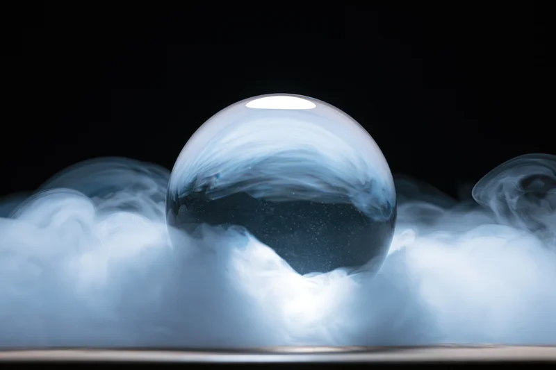 A clear glass crystal ball resting on a flat surface surrounded by swirling smoke against a dark background