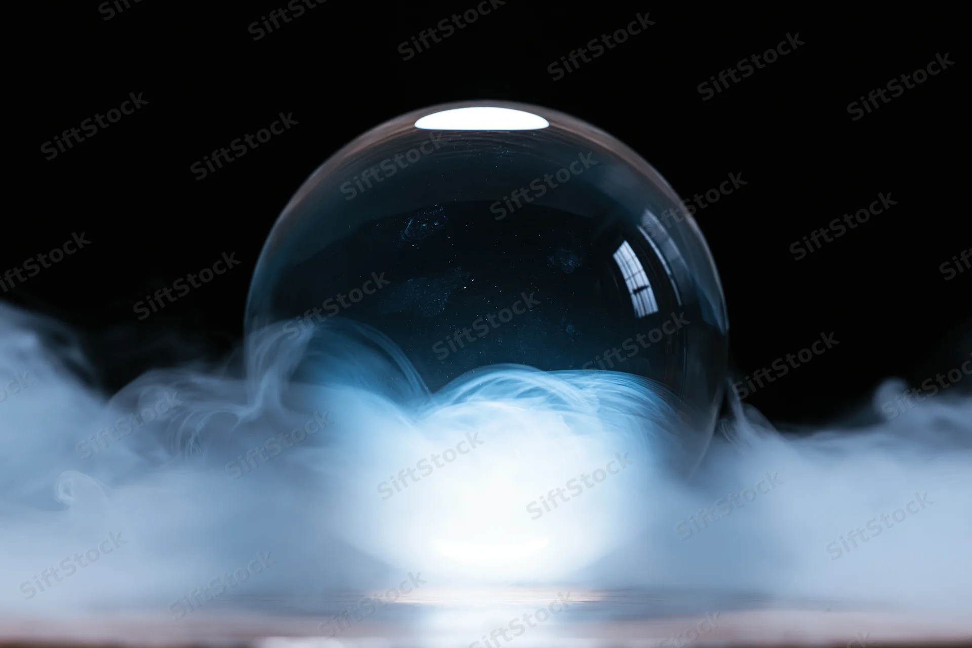 A clear glass crystal ball resting on a surface surrounded by swirling smoke in a dark studio setting