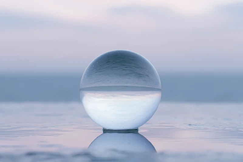 Clear glass lens ball resting on calm wet surface reflecting sky and water