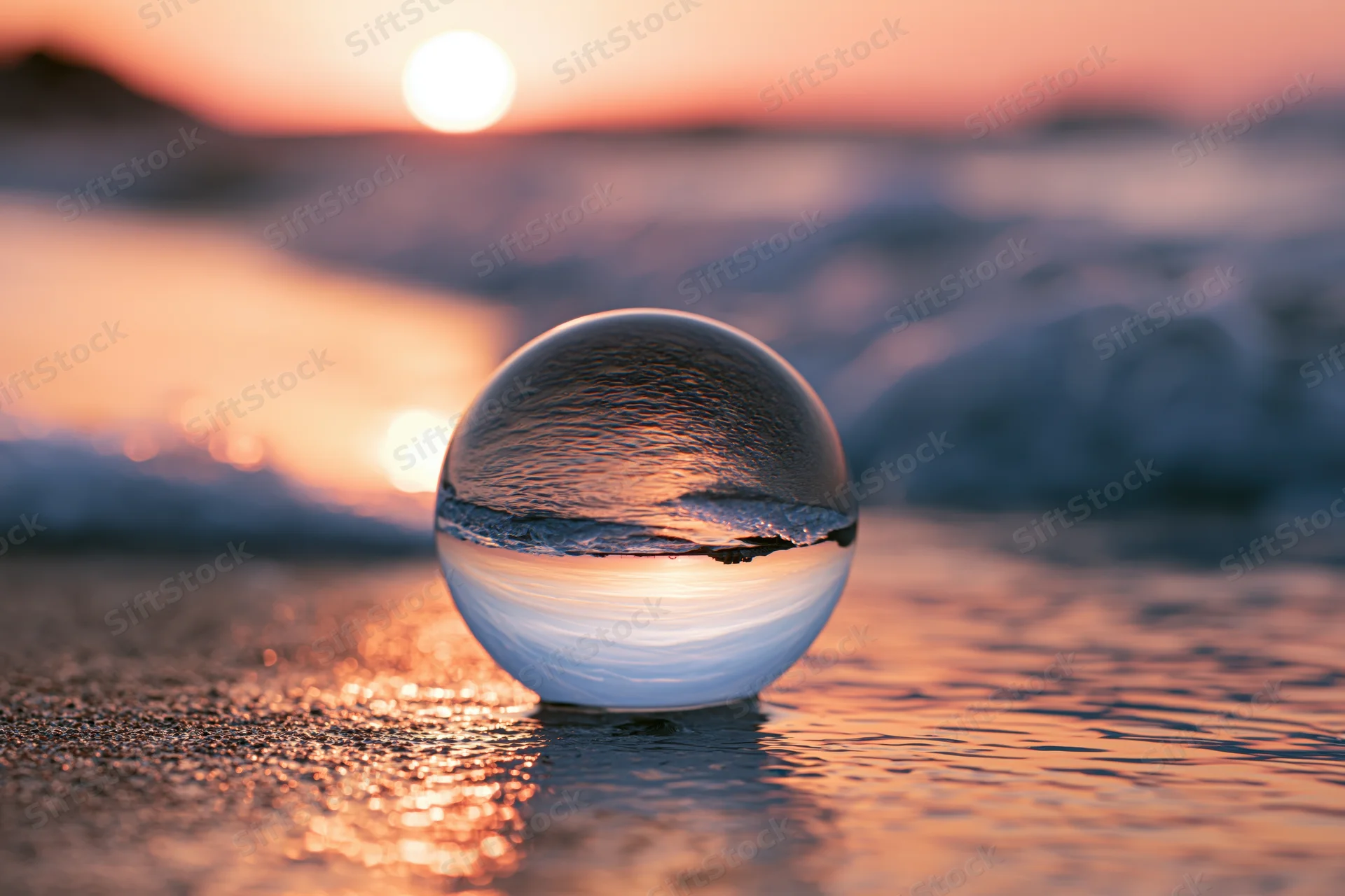 Clear glass lens ball resting on wet sand at ocean's edge during sunset