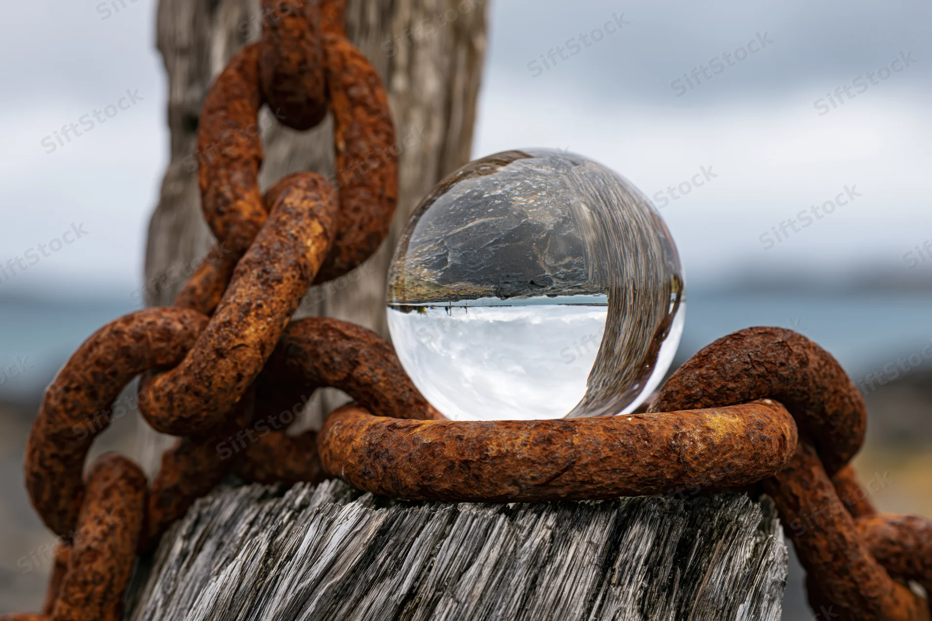 Clear glass lensball resting on rusted iron chain wrapped around weathered wooden post by the ocean