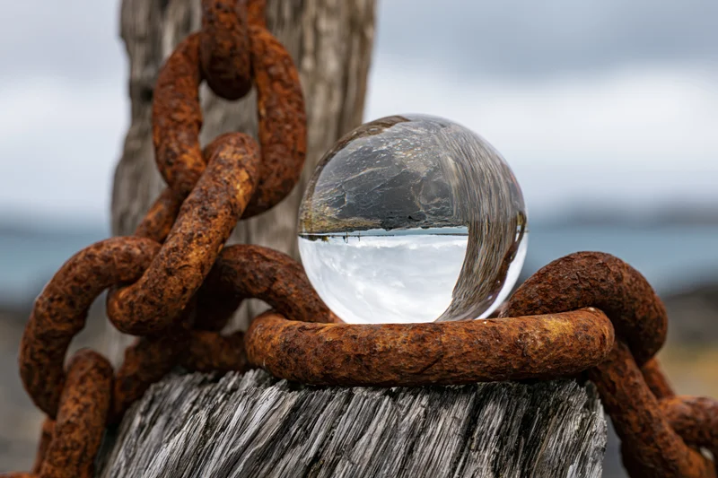 Clear glass lensball resting on rusted iron chain wrapped around weathered wooden post by the ocean