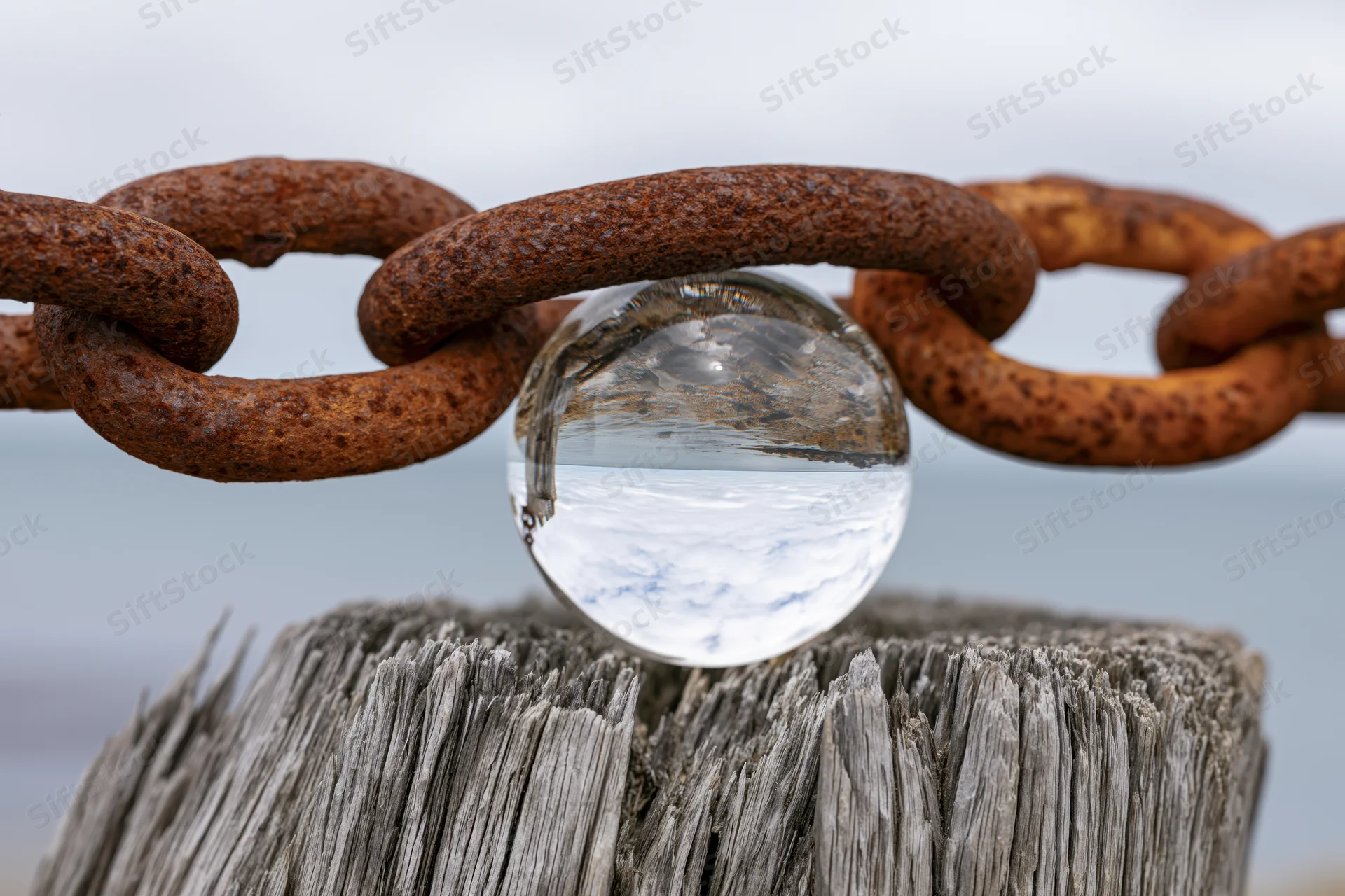 Clear glass lensball resting on rusted iron chain over weathered wooden stump by the ocean
