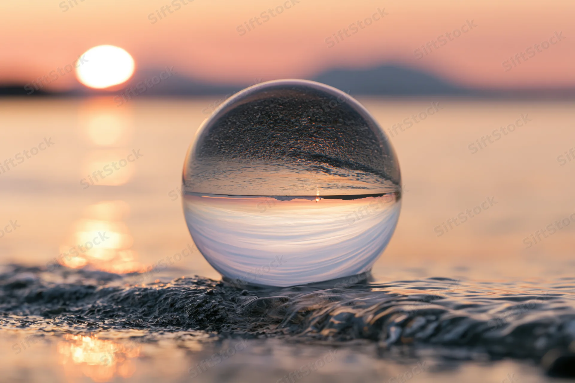 Glass lensball resting on wet sand at beach reflecting sunset over ocean