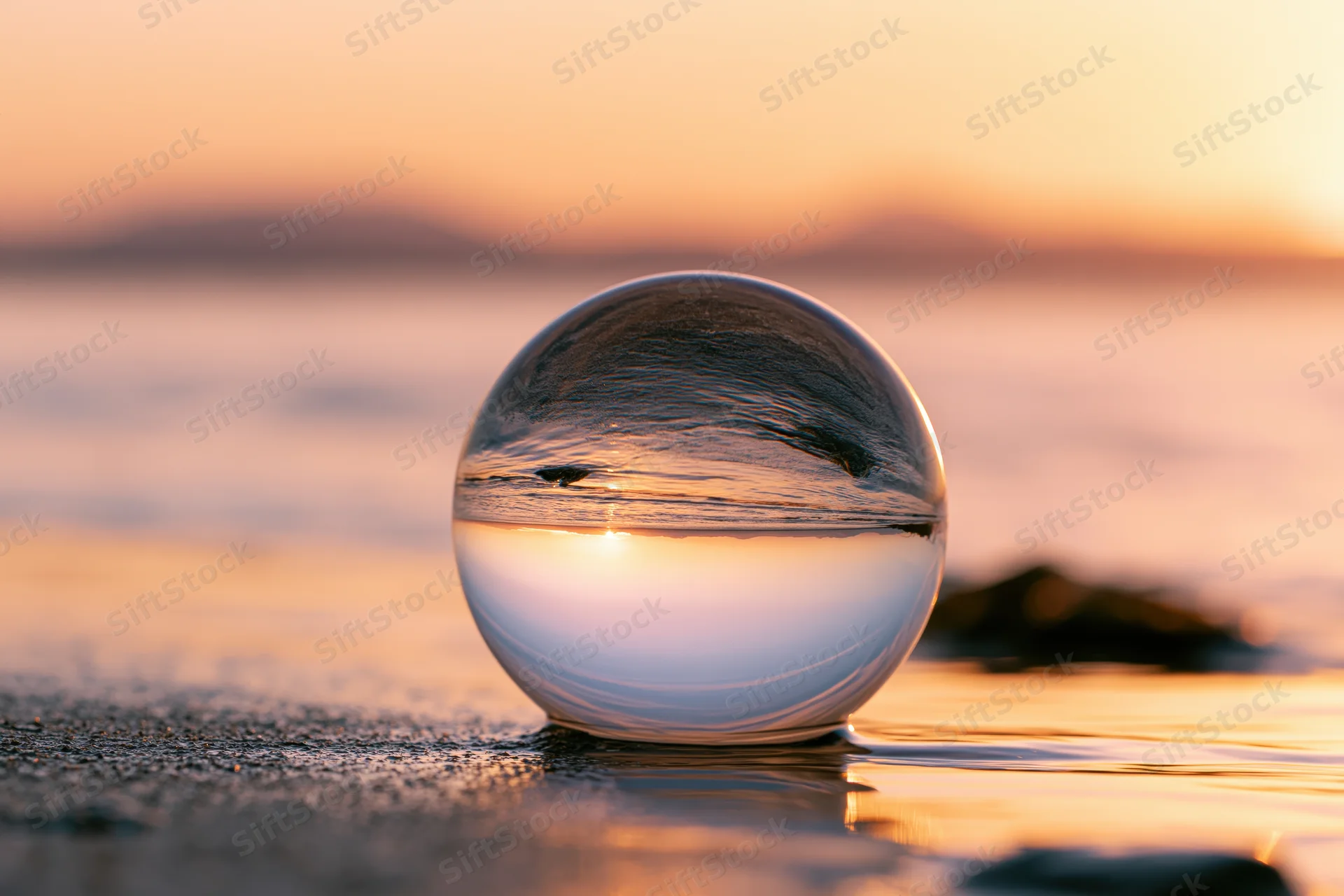 Clear glass lensball resting on wet sand at beach reflecting sunset over ocean