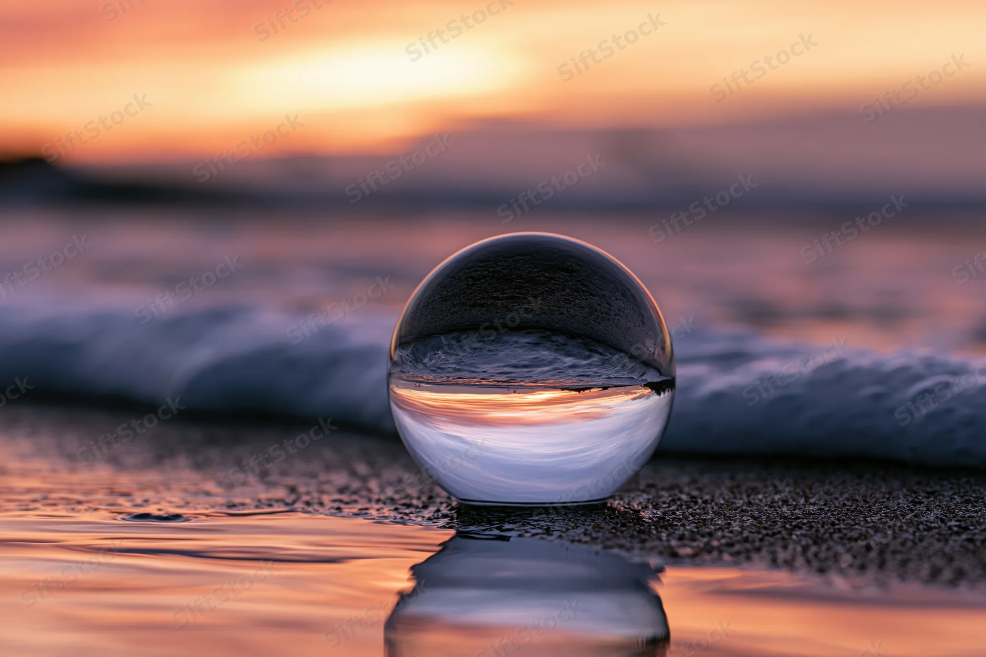 Glass lensball resting on wet sand at ocean's edge during sunset