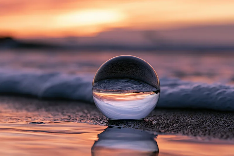 Glass lensball resting on wet sand at ocean's edge during sunset