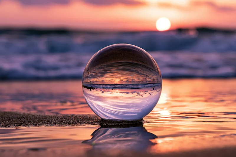 Clear glass lensball resting on wet sand at ocean's edge during sunset