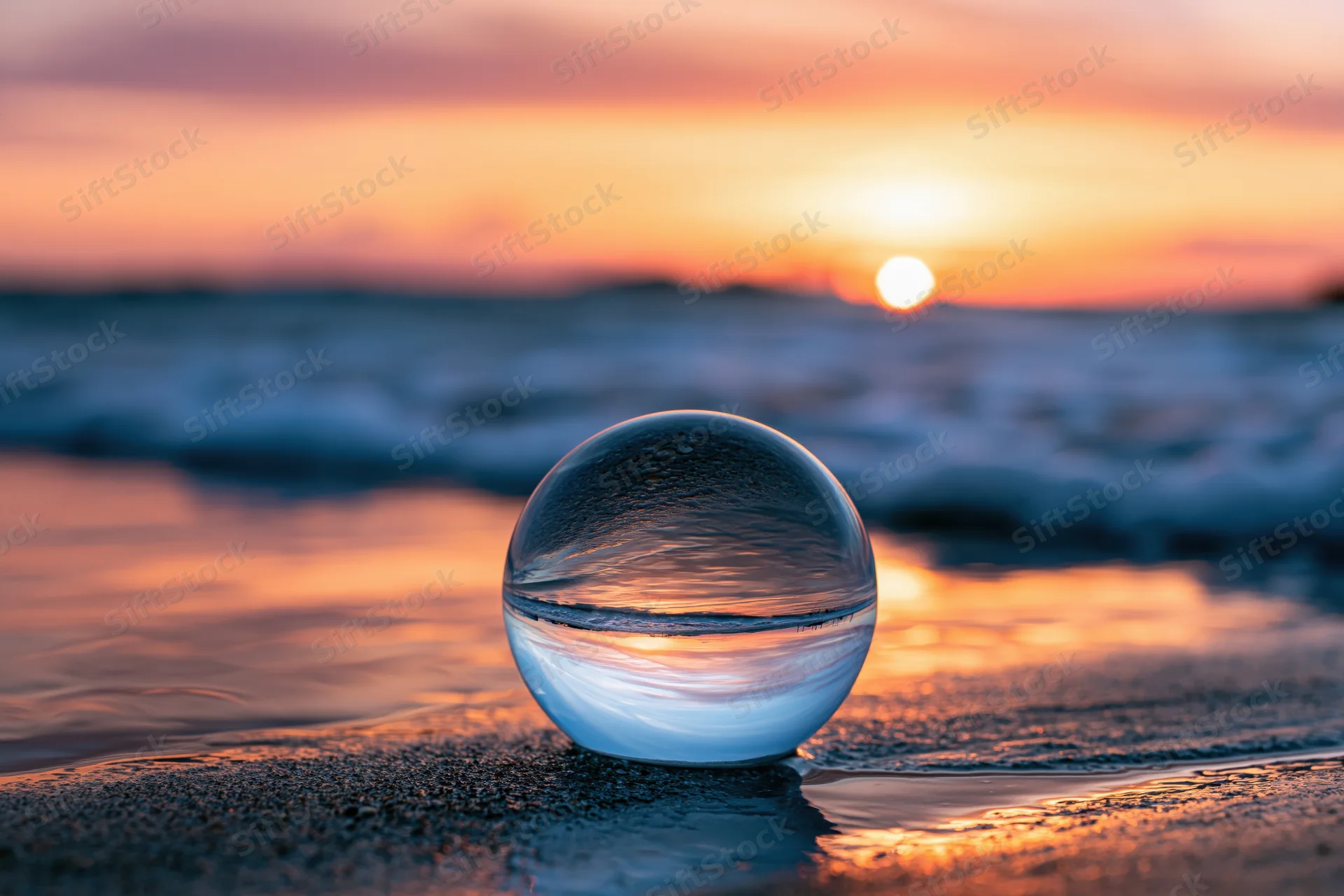 Clear glass lensball resting on wet sand at ocean's edge during sunset