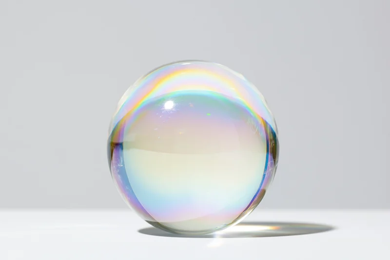 A clear glass sphere with iridescent rainbow reflections sitting on a white surface in a studio setting