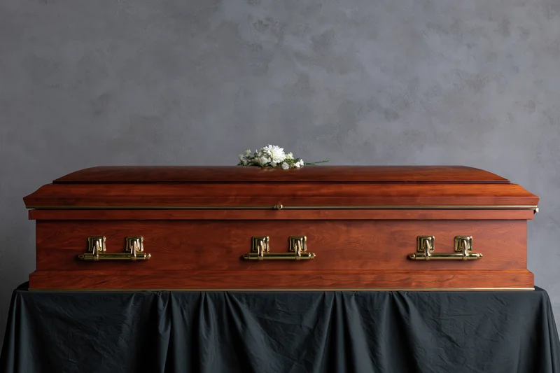 Closed wooden casket with brass handles and white flowers on top, resting on a black draped table against a gray wall