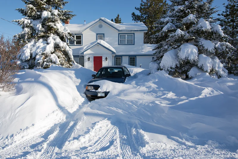 Dark blue sedan buried in deep snow drifts in front of a white house with red door and snow-covered evergreen trees