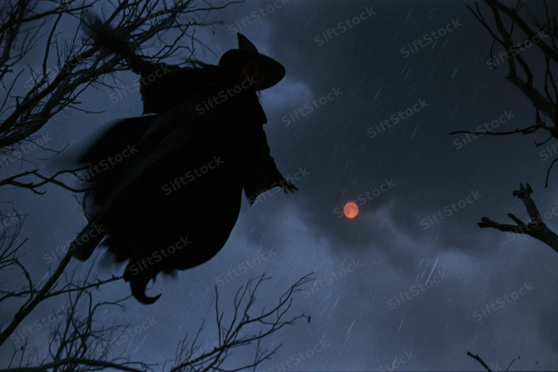 Witch flying on broomstick through rainy night sky with red moon and bare trees