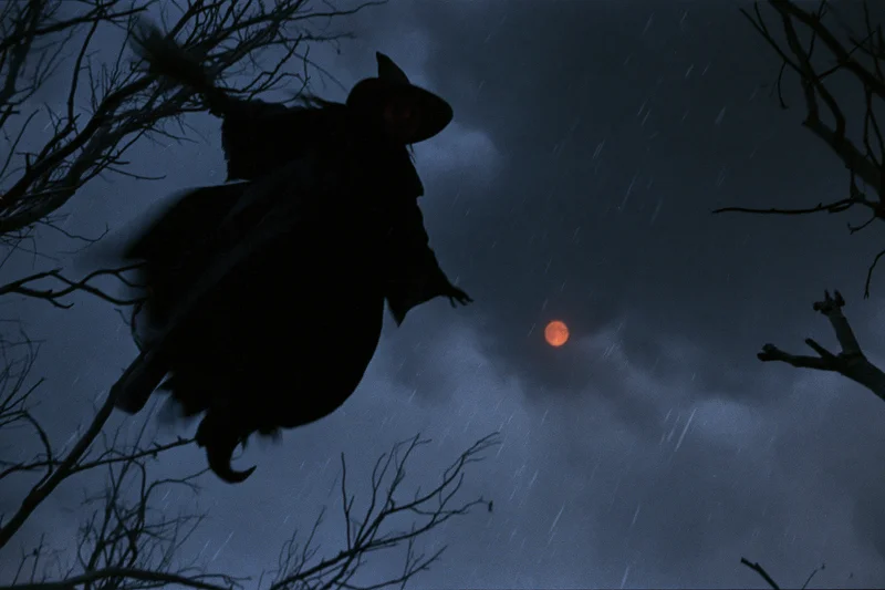 Witch flying on broomstick through rainy night sky with red moon and bare trees