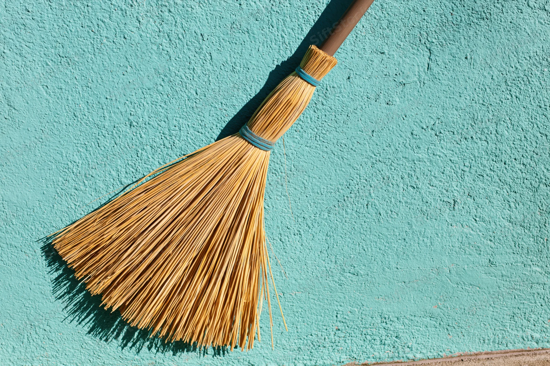 Straw broom leaning upright against a textured blue wall in sunlight