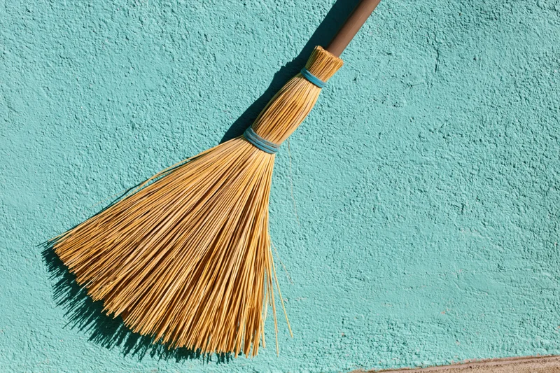 Straw broom leaning upright against a textured blue wall in sunlight