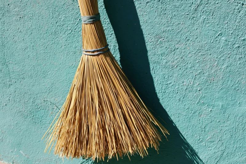 Straw broom leaning upright against a textured turquoise wall in sunlight