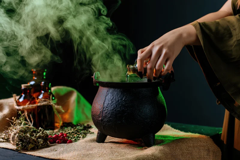 Witch pouring liquid into a smoking cauldron on a table with herbs and bottles