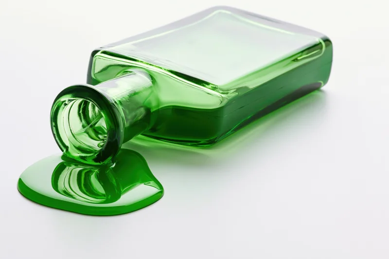 Green glass bottle lying on its side with spilled liquid on white surface