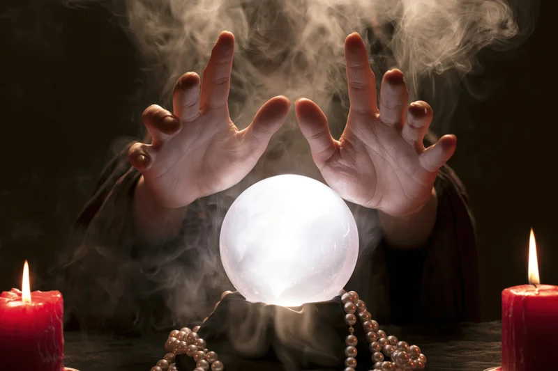 Hands hovering over a glowing crystal ball surrounded by candles and beads on a dark table